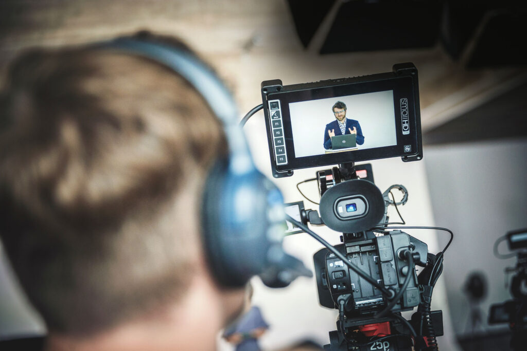 Nahaufnahme einer Videokamera, die eine moderierende Person auf dem Display zeigt, während ein Kameramann mit Headset filmt
