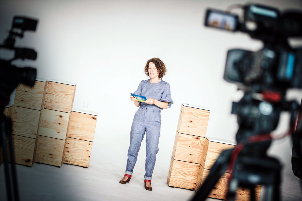 Moderatorin steht zwischen hölzernen Boxen in einem hellen Studio und liest von einer Karte, während Kameras sie aufnehmen